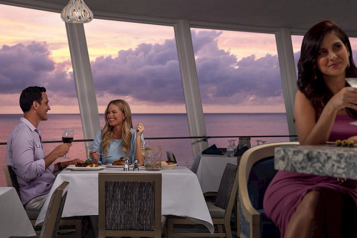 A couple is dining by the sea at sunset while a woman in the foreground enjoys a cocktail.