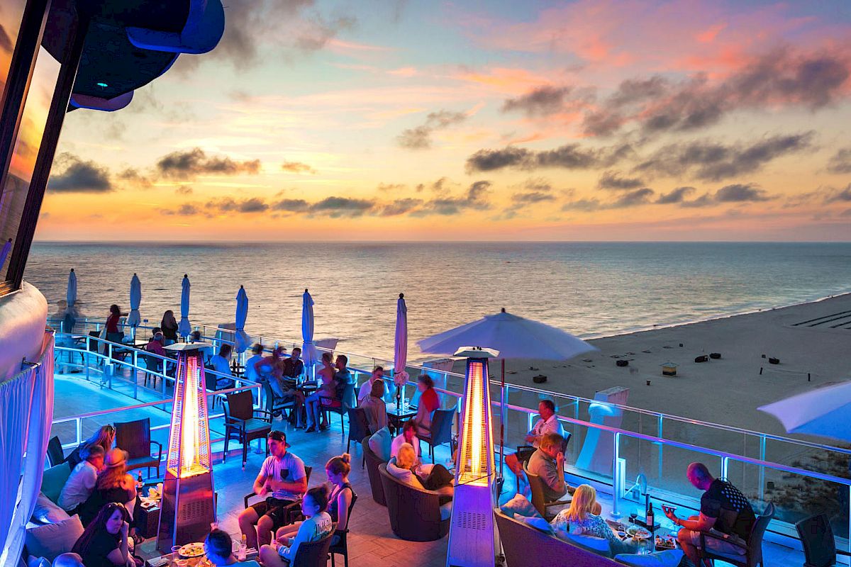 People enjoying an evening at a beachside restaurant or bar, with a stunning sunset view over the ocean and blue lighting.