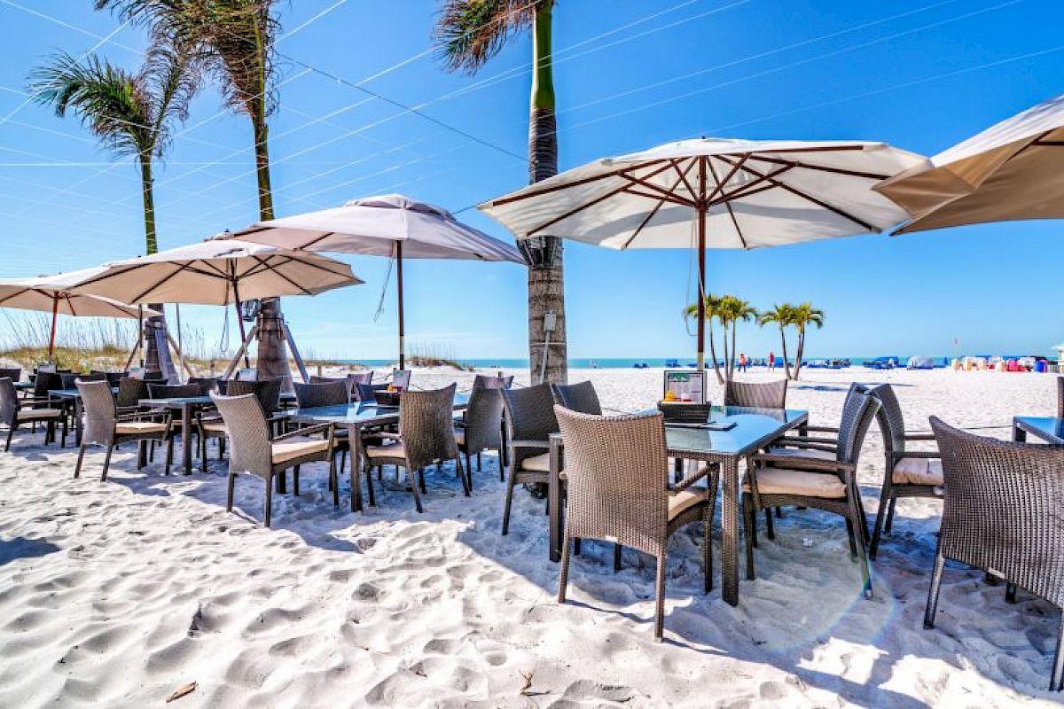 Outdoor beachside restaurant with tables and chairs under large umbrellas on white sand, with palm trees and blue sky in the background.