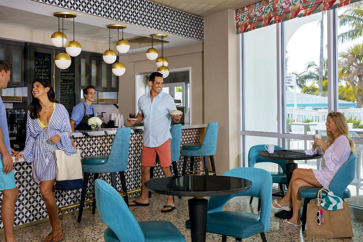 People socializing in a stylish café with teal and black decor, modern lights, and a view of palm trees through large windows.
