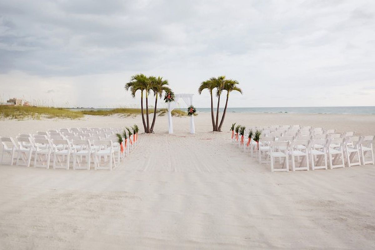 Beach wedding setup with white chairs and palm trees awaits guests.