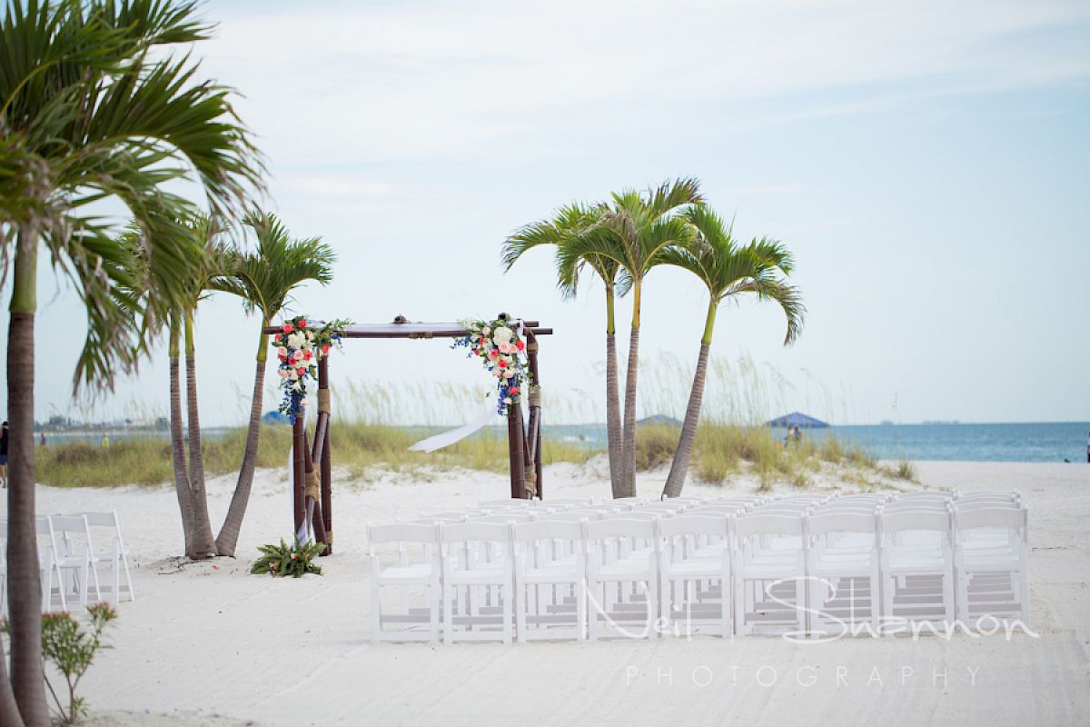 Beach wedding setup with decorated arch, white chairs and palm trees.