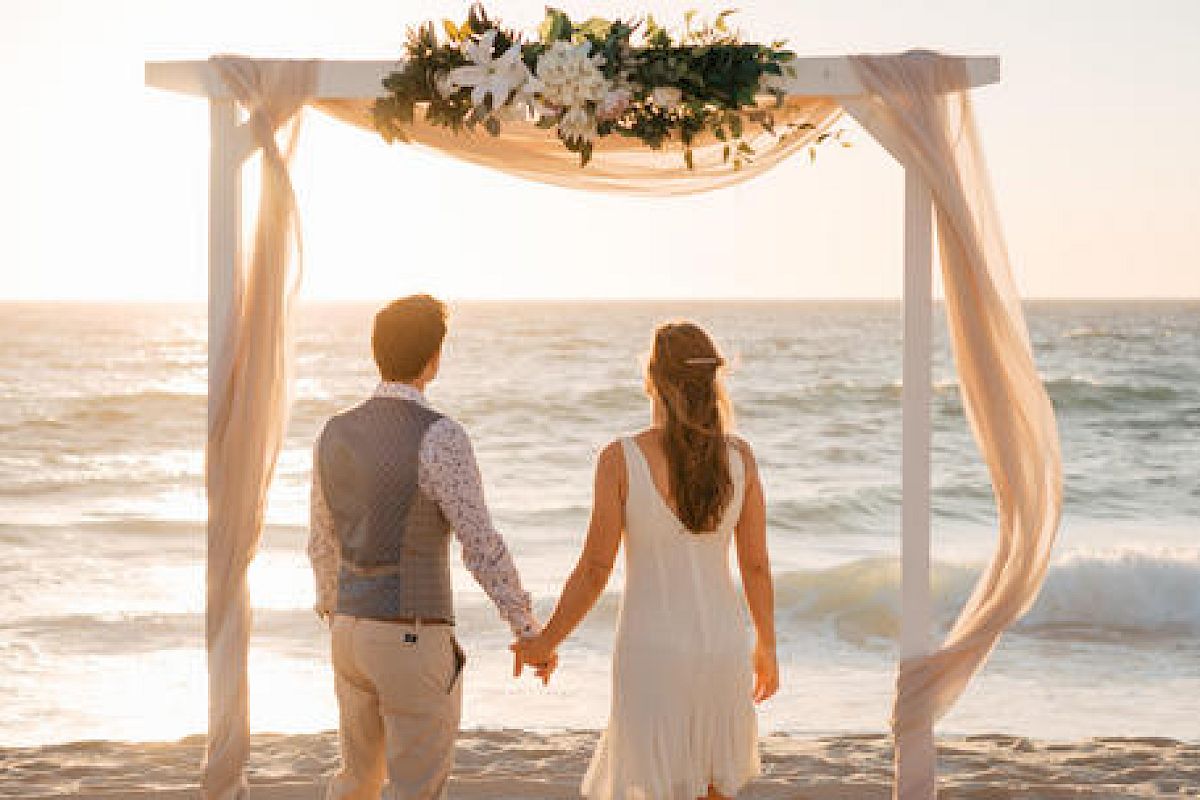 A couple holds hands under a floral arch by the sea at sunset.