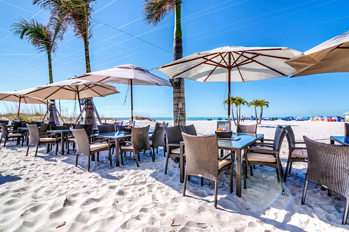 A beachside outdoor dining area with tables, chairs, and umbrellas on white sand.