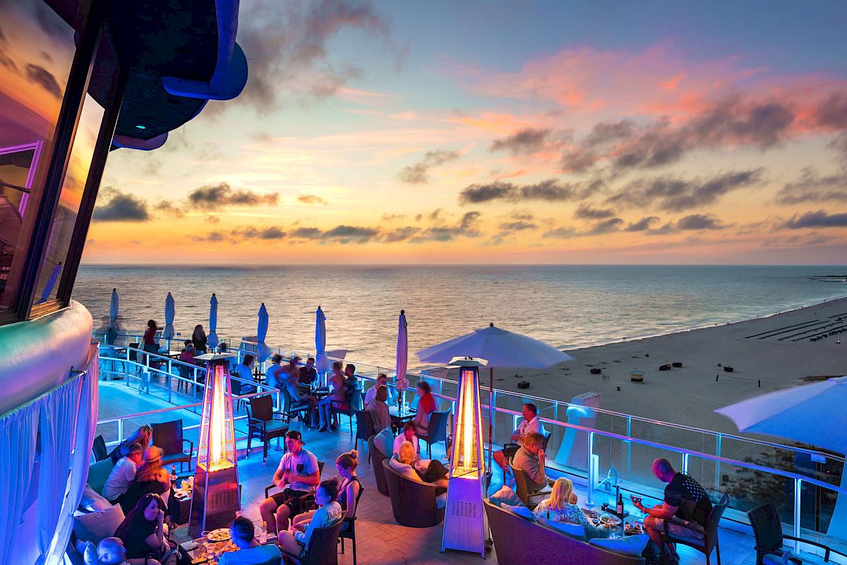 A seaside rooftop bar with people dining, ambient lighting, and a colorful sunset sky.
