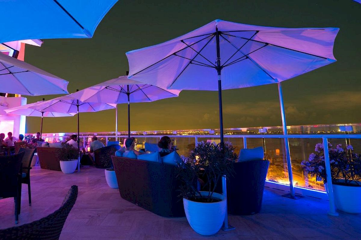 Rooftop bar at night with umbrellas, people sitting, and a view of the city lights.