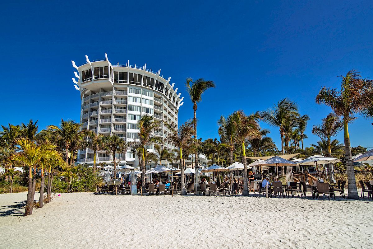 A sunny beachfront with palm trees, a chic outdoor cafe, and a unique high-rise building.