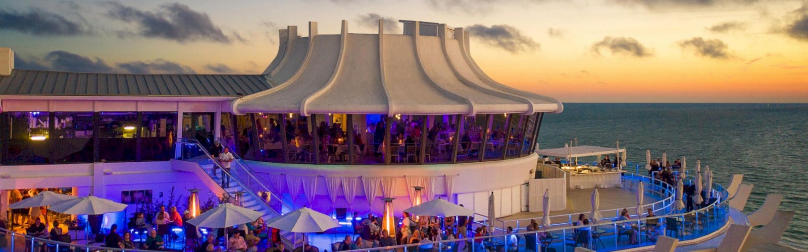 A rooftop bar at sunset with people, seating areas, and a lit-up bar.