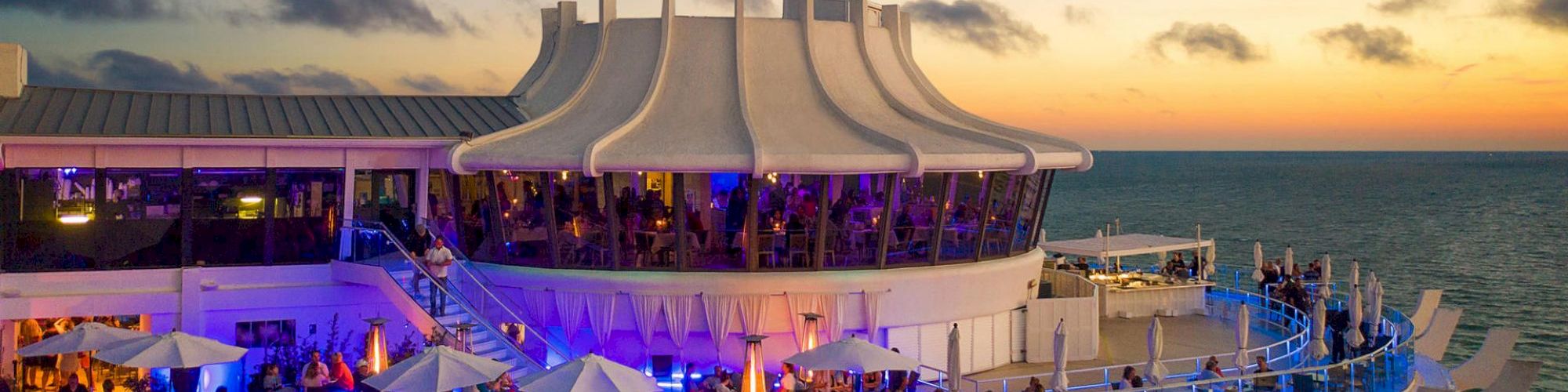 A cruise ship deck at sunset with people, seating areas, and a lit-up bar.