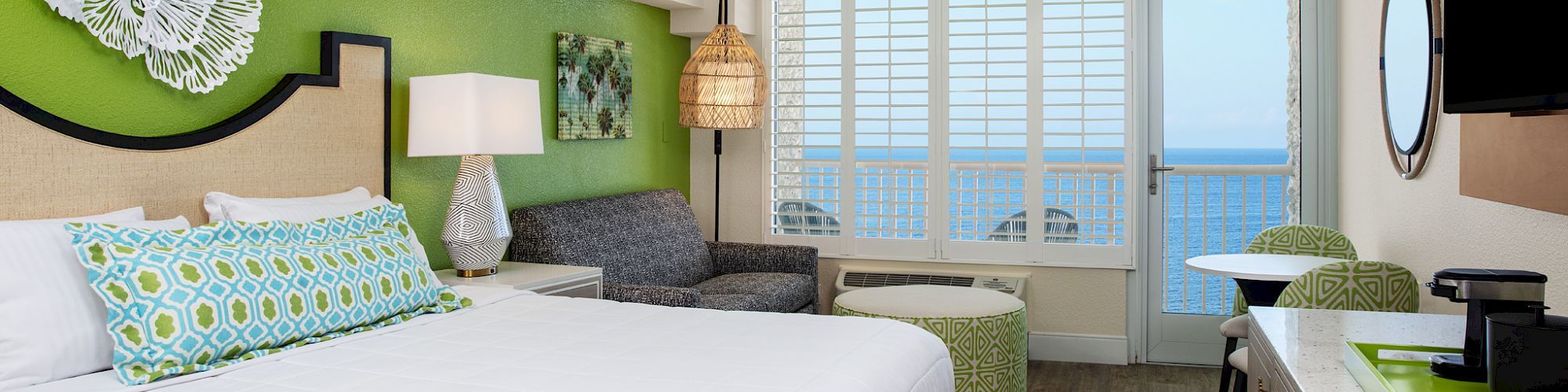 A modern hotel room with a bed, ocean view, seating area, and bright decor.