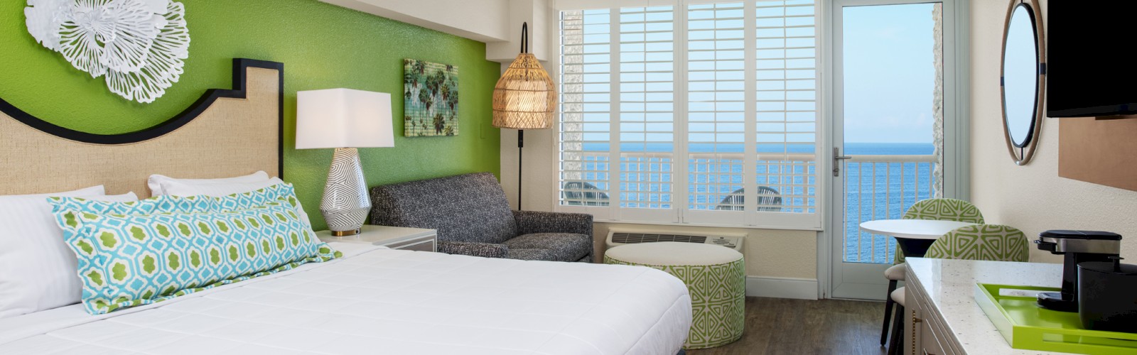 A modern hotel room with a bed, ocean view, seating area, and bright decor.