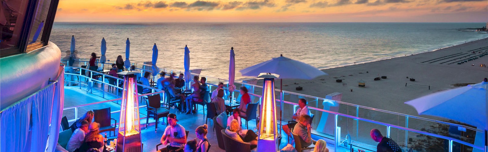 People are enjoying a rooftop patio with tables, heaters, and umbrellas, overlooking a beach at sunset with a colorful sky.