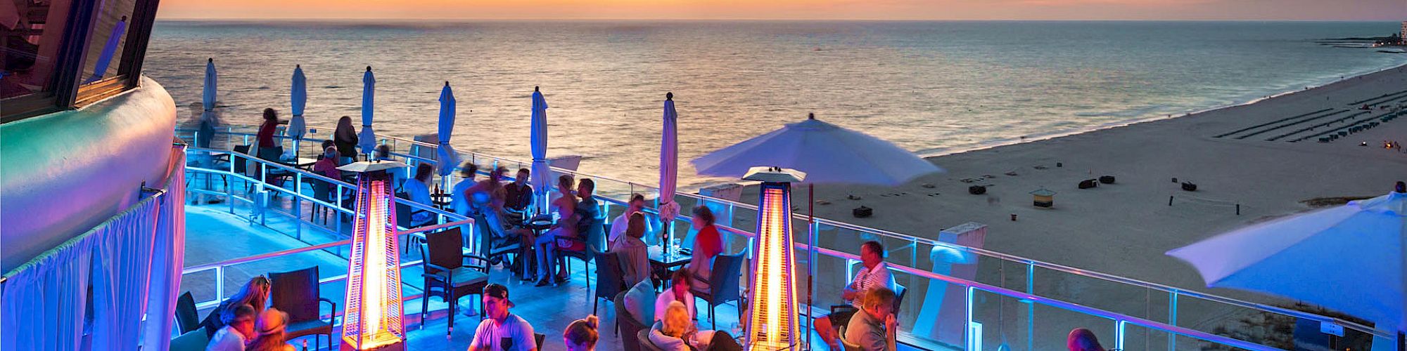 People are enjoying a rooftop patio with tables, heaters, and umbrellas, overlooking a beach at sunset with a colorful sky.