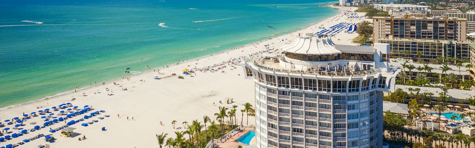 Aerial view of a beach, ocean, building with a unique roof, umbrellas, and palm trees.