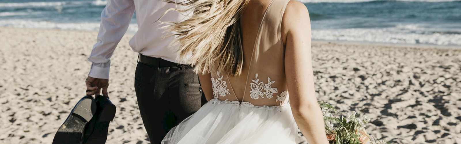 A couple holding hands, likely newlyweds, on a beach with waves in the background.