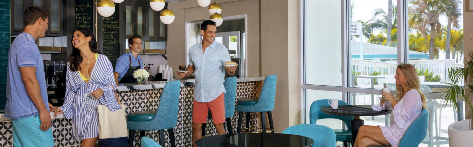 People are inside a chic café with modern decor. Some are at the counter ordering beverages, while others are sitting at tables, enjoying their drinks.