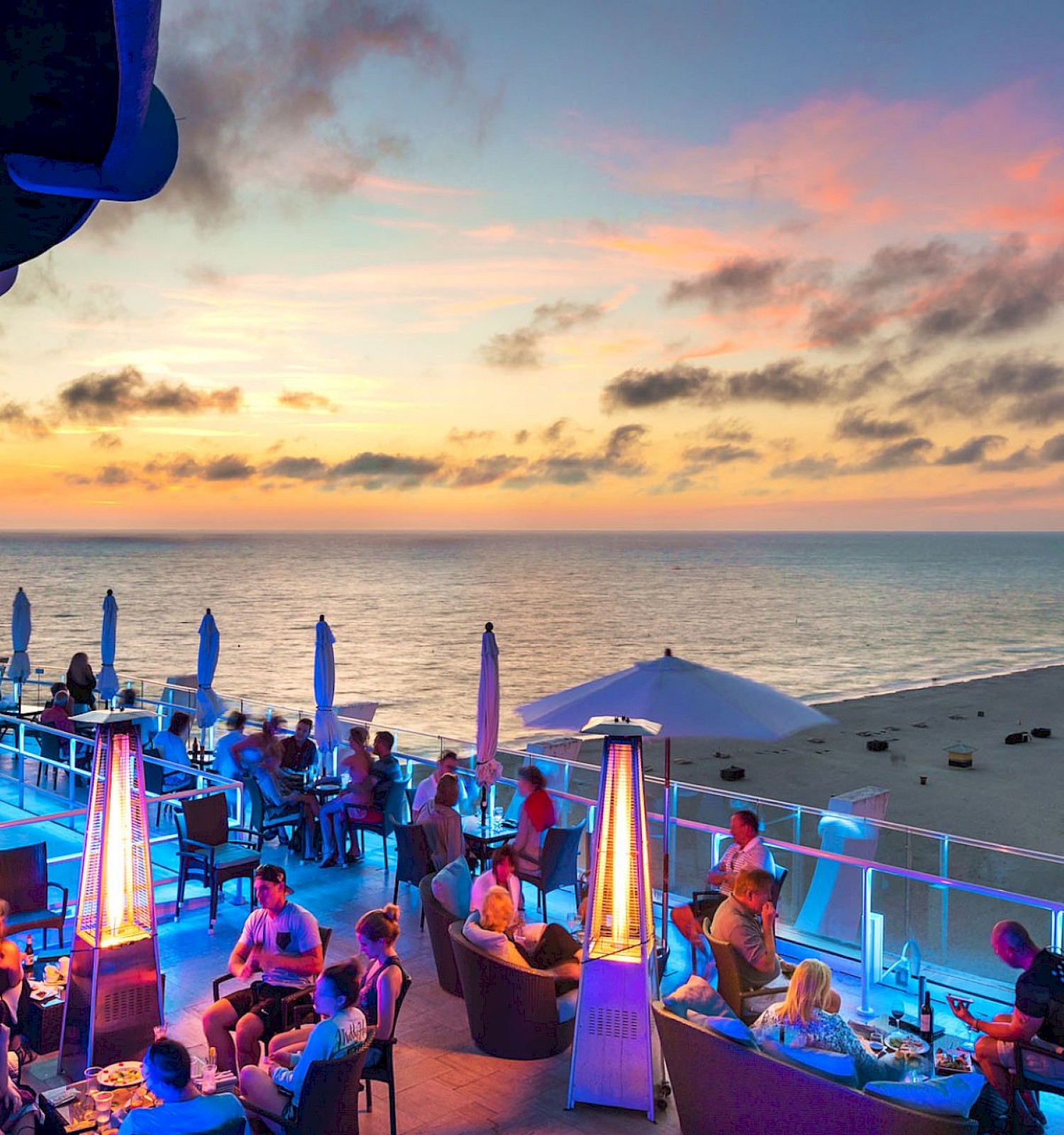 People relax at a beachfront restaurant with outdoor seating, heaters, and sunset views over the ocean.