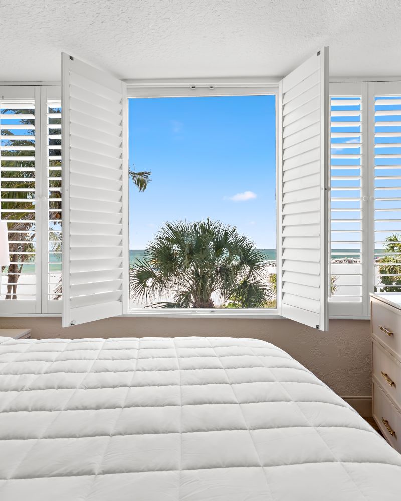 A bright bedroom with an open window showing palm trees, a bed with blue stripes, a dresser, and a mounted TV.