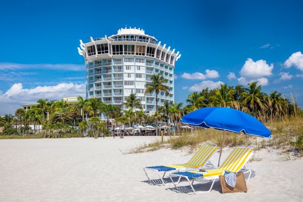 Beach with a blue umbrella, two lounge chairs, palm trees, and a unique round building under the blue sky.