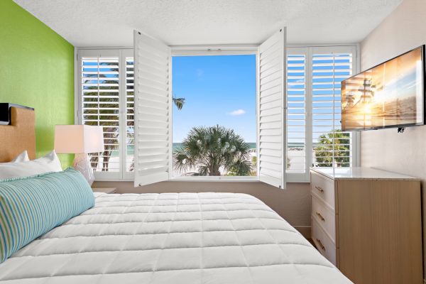 Bright bedroom with an open window looking out to palm trees, a bed in the foreground, and a mounted TV.