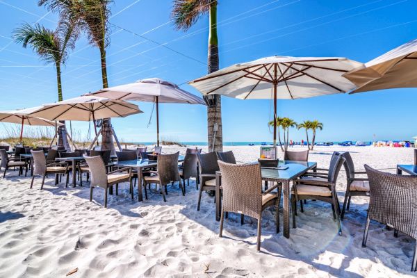 Outdoor beachside dining area with tables, chairs, umbrellas, and palm trees on sandy ground.