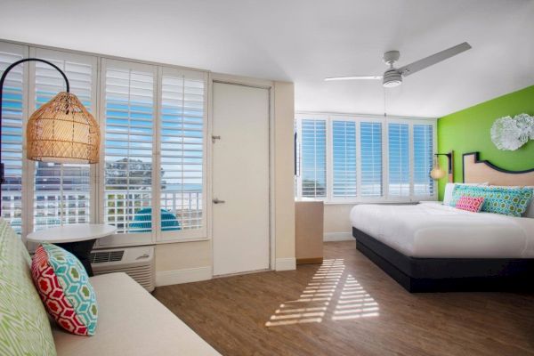 Bright hotel room with a large bed, colorful pillows, a ceiling fan, and ocean view.