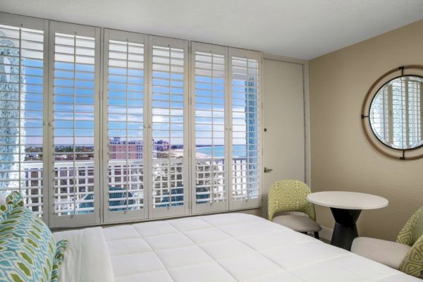 A room with a large bed, window blinds open to city view, chairs, table, and a decorative mirror.