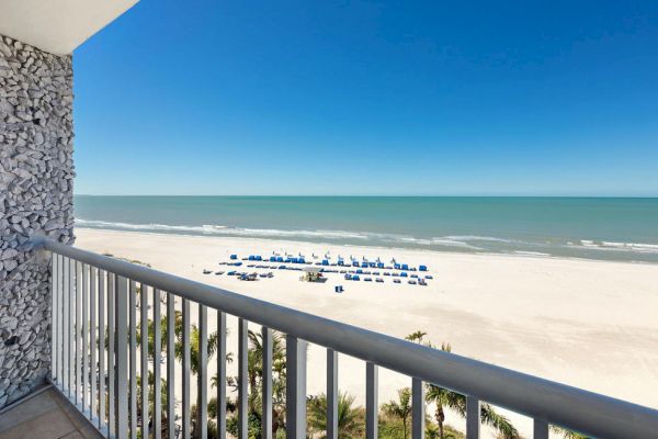 A balcony view of a beach with blue umbrellas and chairs, clear sky, and calm sea.