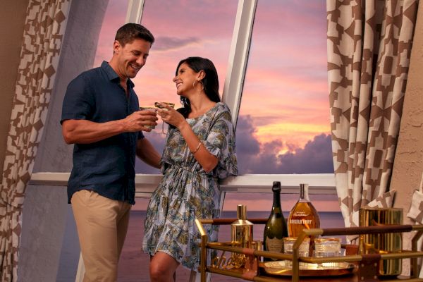 A couple is toasting drinks at sunset on a balcony, with a wine setup nearby.