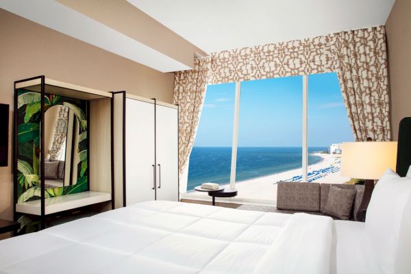 Modern room with large window overlooking the beach, white bed, and decorative mirror.