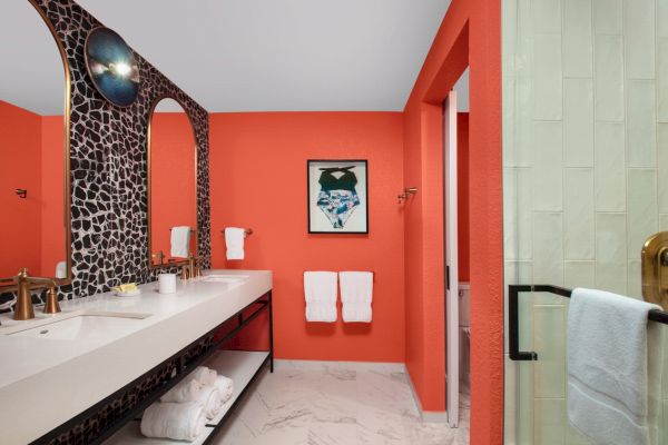 A vibrant orange bathroom with a white sink, towels, and a uniquely shaped mirror.