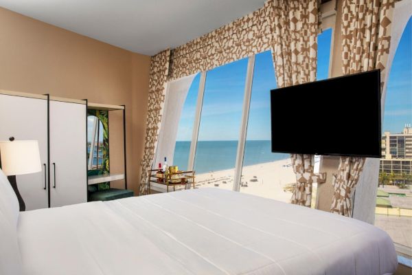 A hotel room with a bed, TV, and a view of the beach through large windows.