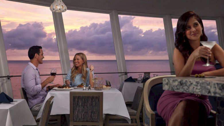 A couple is dining by the sea at sunset while a woman in the foreground enjoys a cocktail.