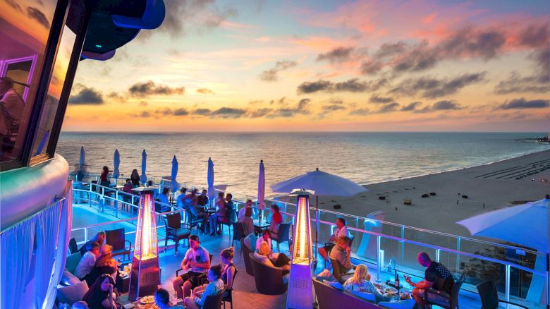People enjoying an evening at a beachside restaurant or bar, with a stunning sunset view over the ocean and blue lighting.