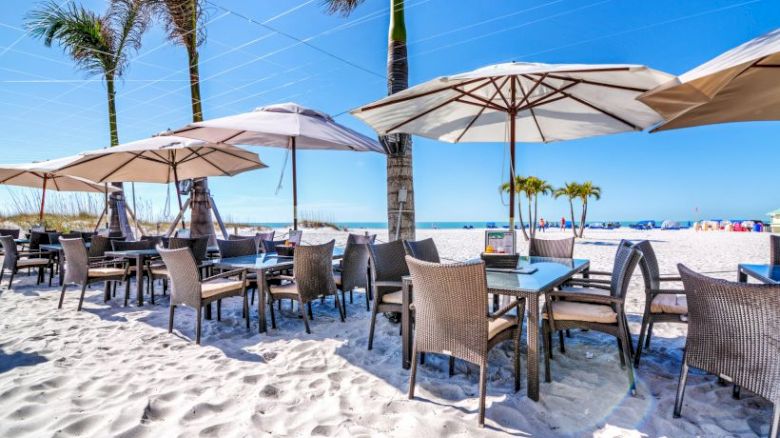 Outdoor beachside restaurant with tables and chairs under large umbrellas on white sand, with palm trees and blue sky in the background.