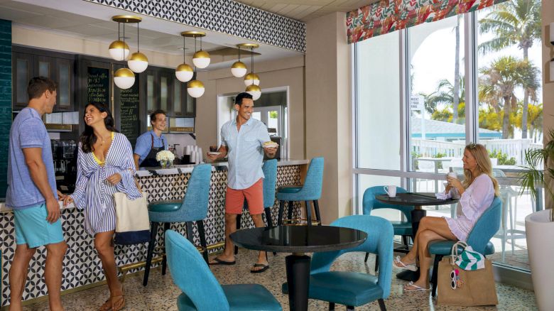 People socializing in a stylish café with teal and black decor, modern lights, and a view of palm trees through large windows.