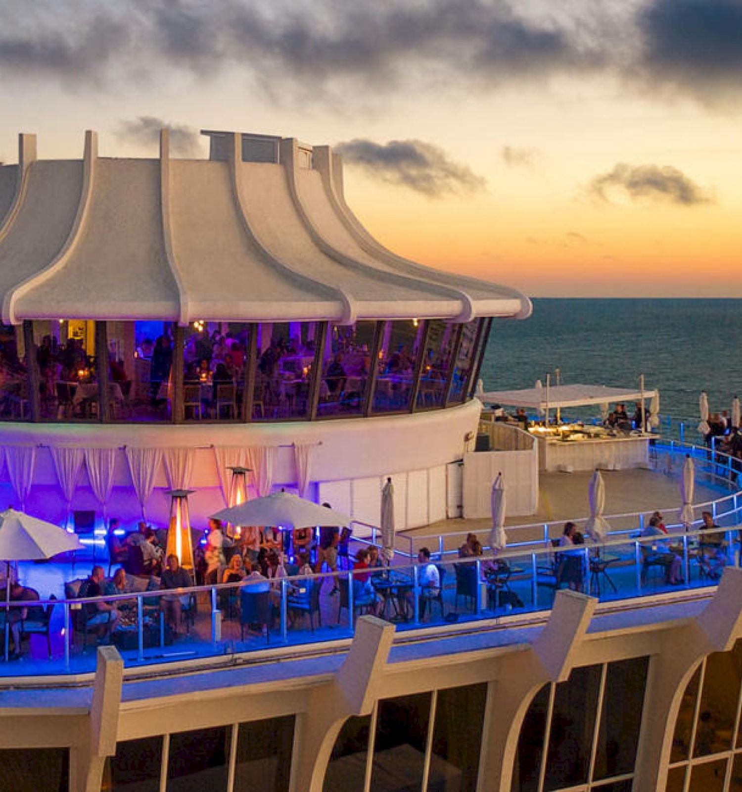 An oceanfront restaurant during sunset with modern architecture, outdoor seating, and a vibrant atmosphere. People are dining and enjoying the view.