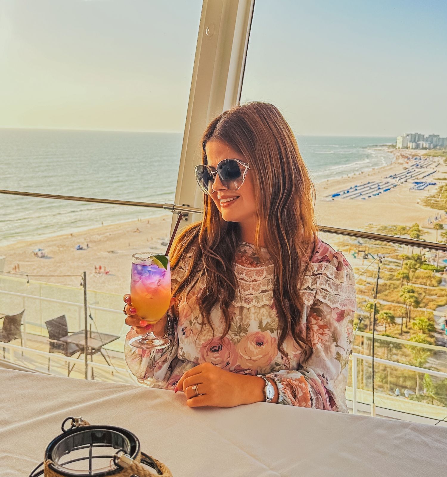 A woman with long hair and sunglasses holds a drink at a beachside table, with a stunning view of the ocean and sandy beach in the background.