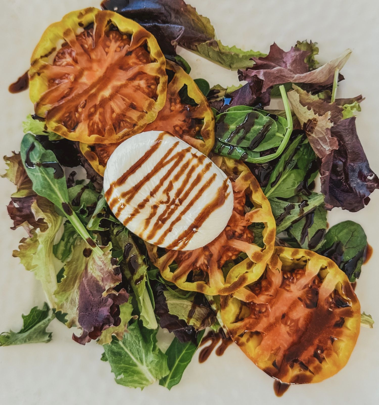 The image shows a salad with mixed greens, three slices of yellow tomato, and a slice of mozzarella cheese, drizzled with balsamic glaze.