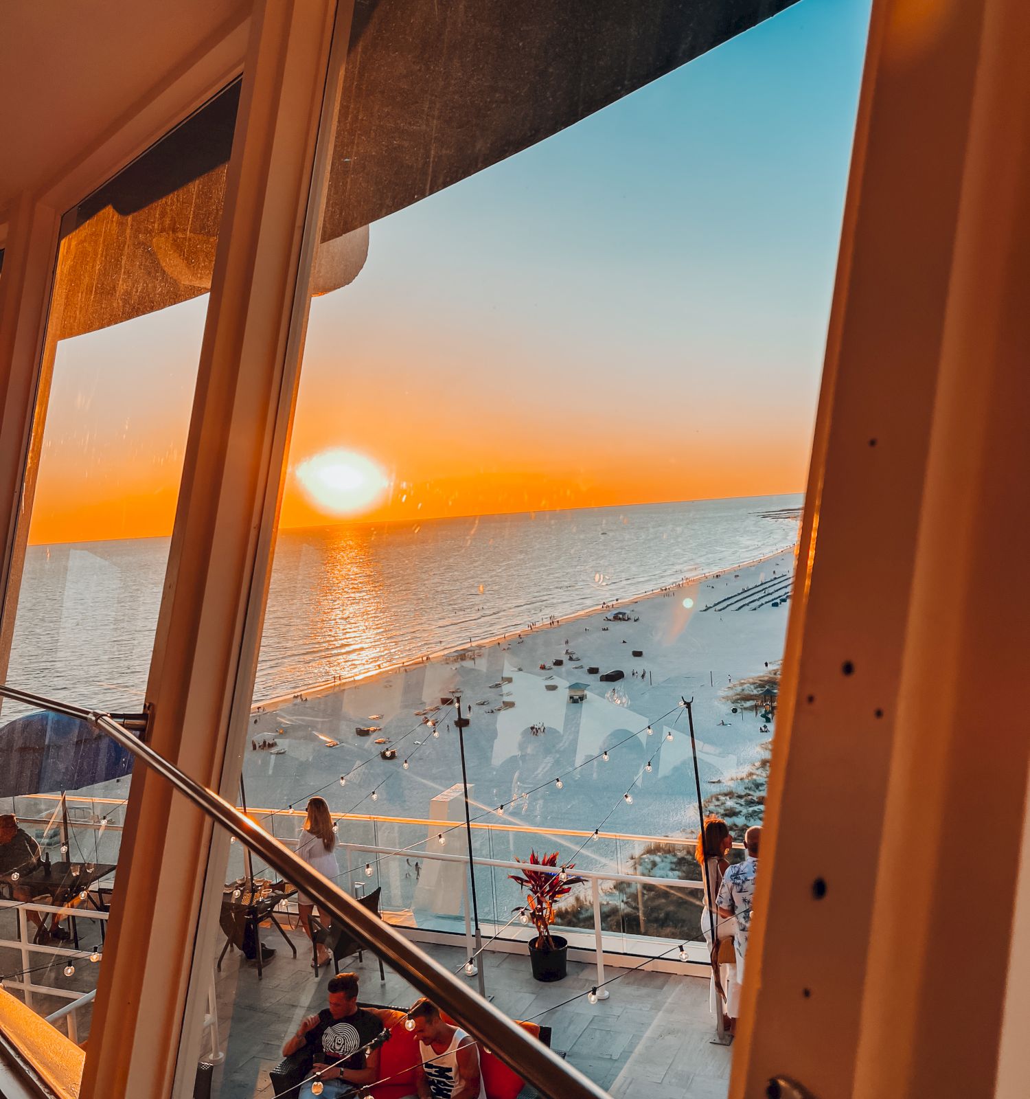 A sunset view over the ocean, seen through a window, with people seated outdoors on a terrace below.