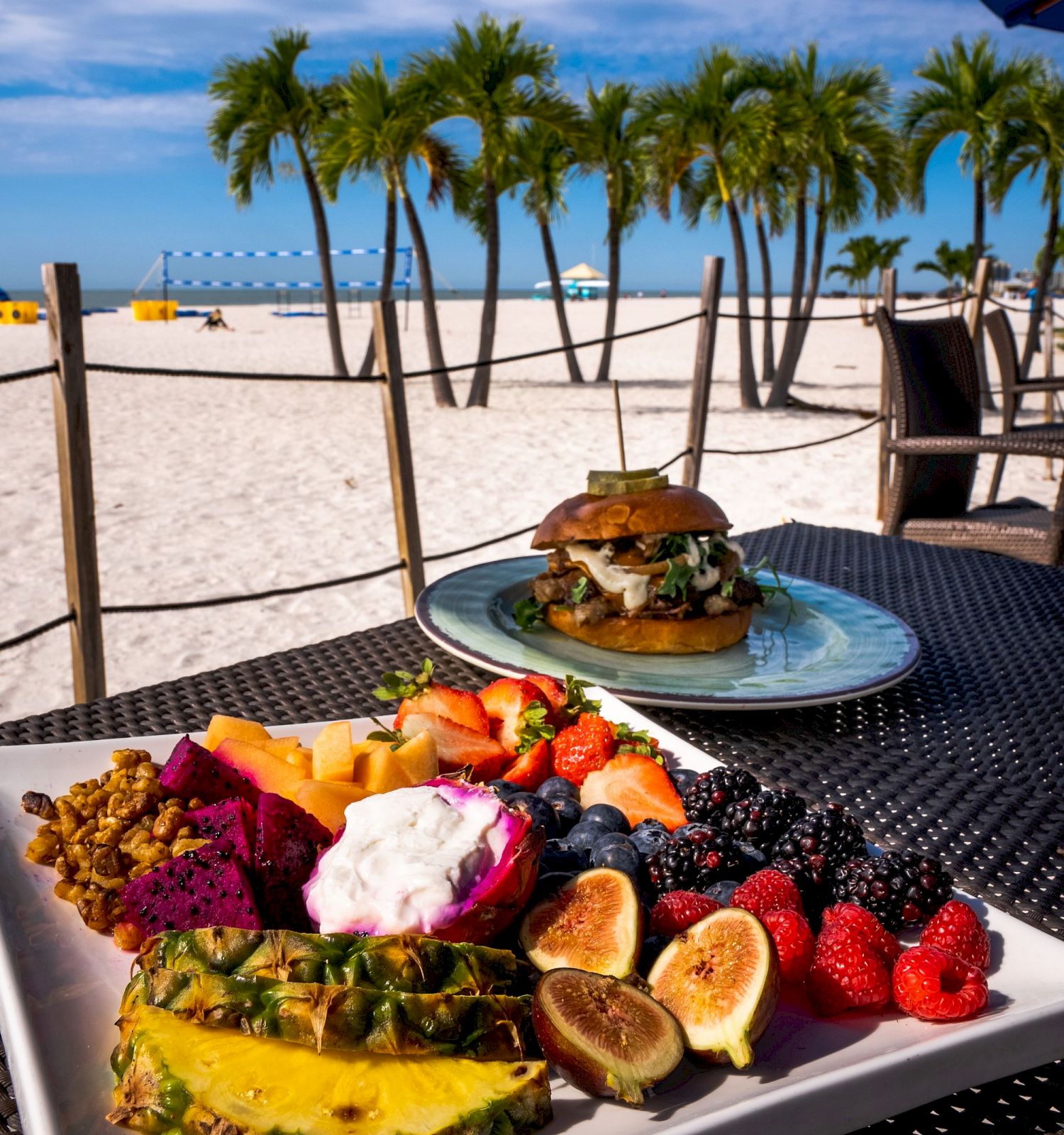 A plate of tropical fruit and a sandwich, on a table by a beach with palm trees and a pier, under a blue sky.