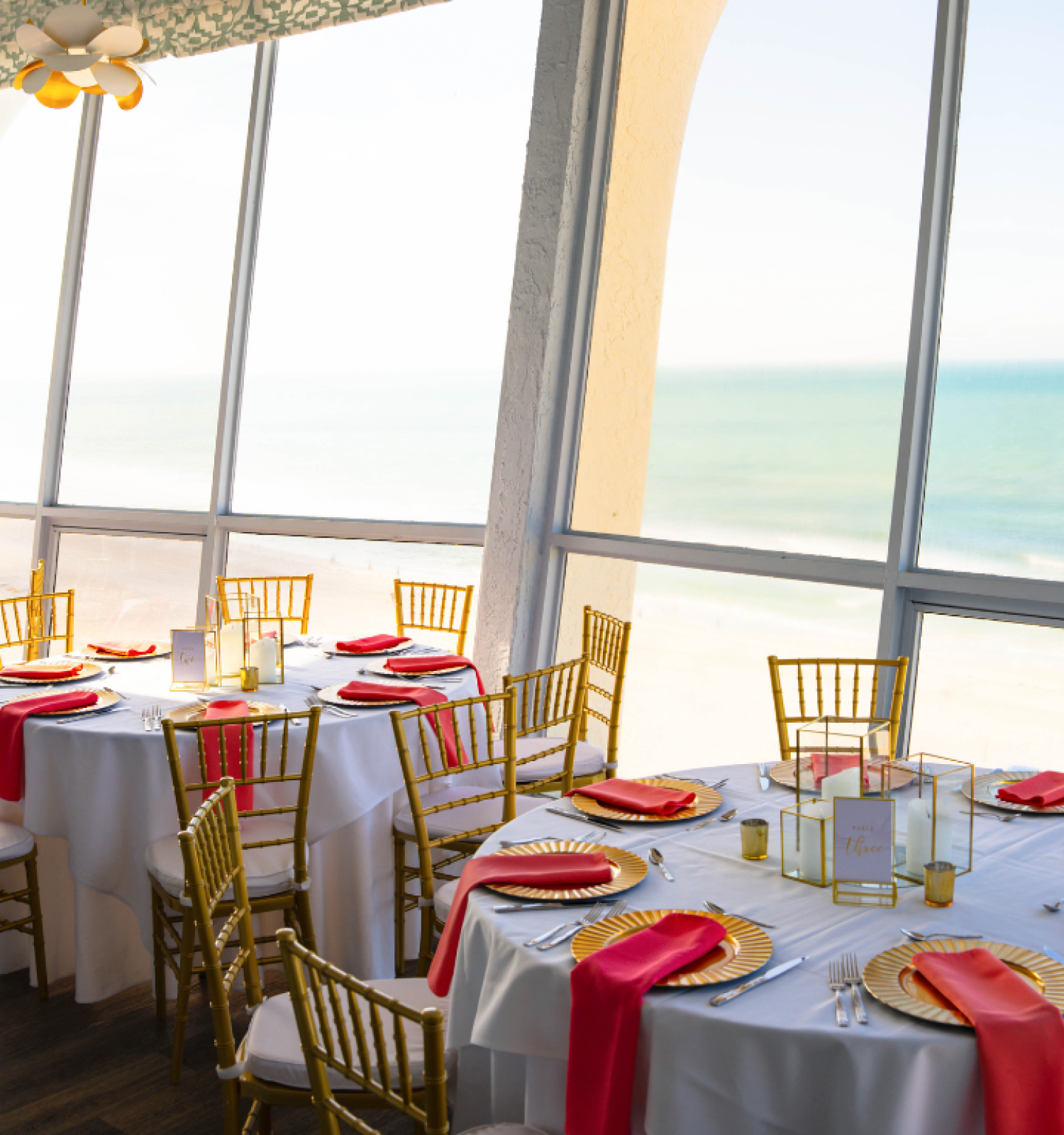 Dining room set for a meal with ocean view, red napkins, and white tablecloths.