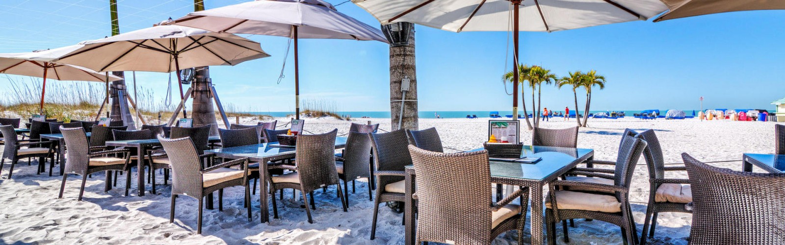 Outdoor beach dining setup with tables, chairs, umbrellas, palm trees, and a bright blue sky.