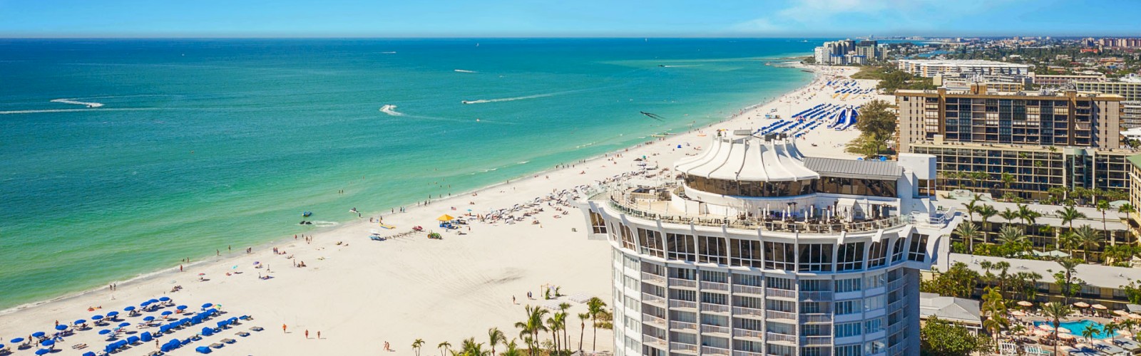 Aerial view of a beachfront with a large building, umbrellas, clear skies, and calm sea.