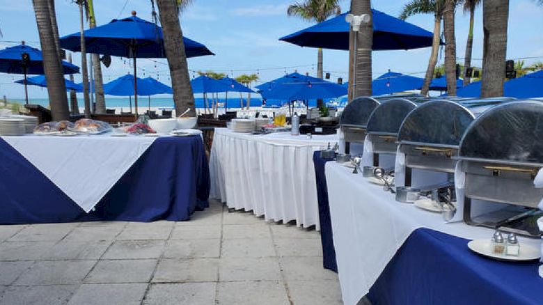 Outdoor buffet setup with blue umbrellas and palm trees in the background.