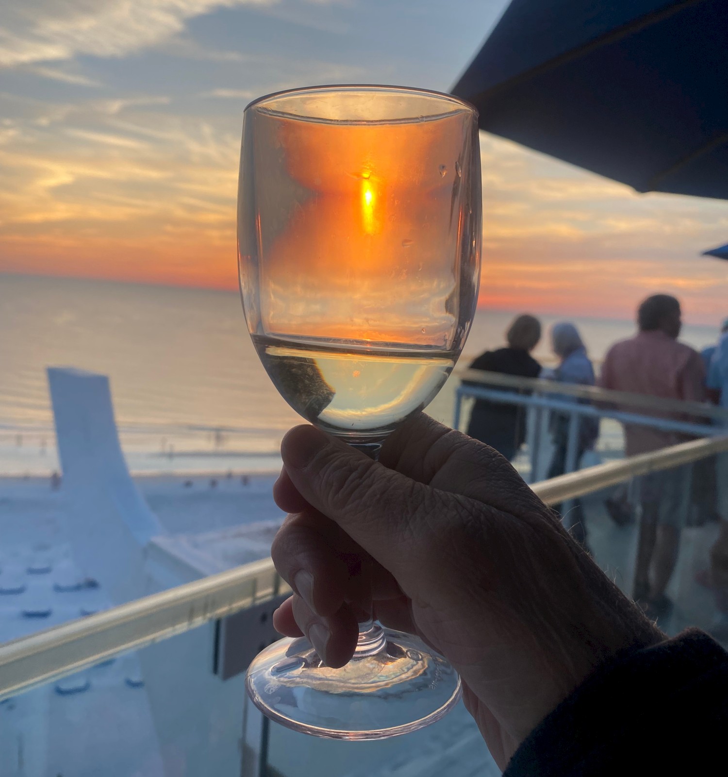 A hand holding a wine glass, with the sunset reflecting through it, and a coastal view with people in the background.