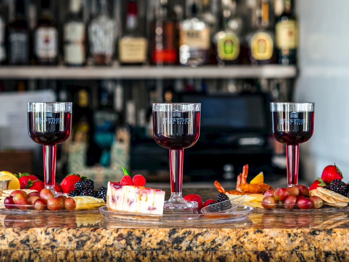Three glasses of red wine are on a bar counter, surrounded by a variety of fruits, cheese, and desserts. Bottles of liquor are in the background.