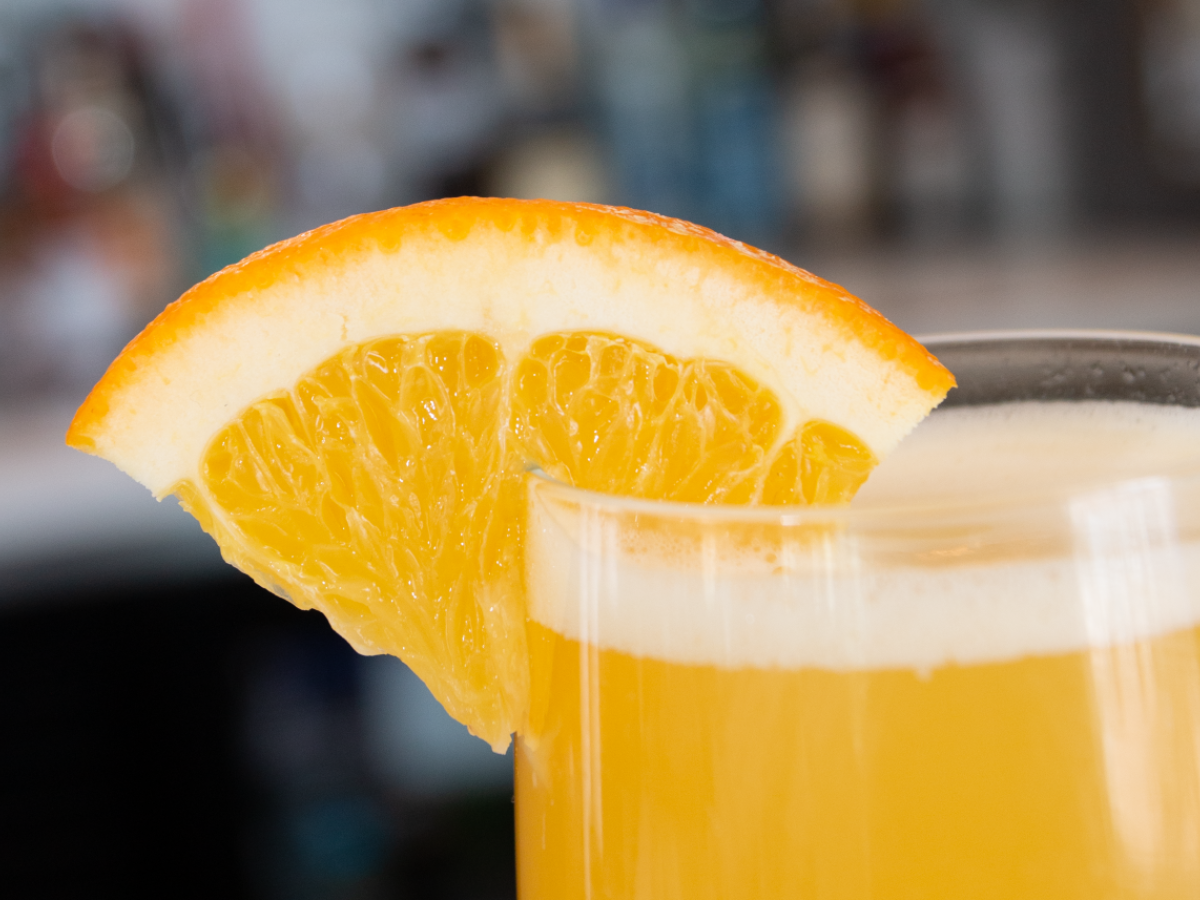 A glass of a yellow-orange beverage garnished with a slice of orange is shown close up.