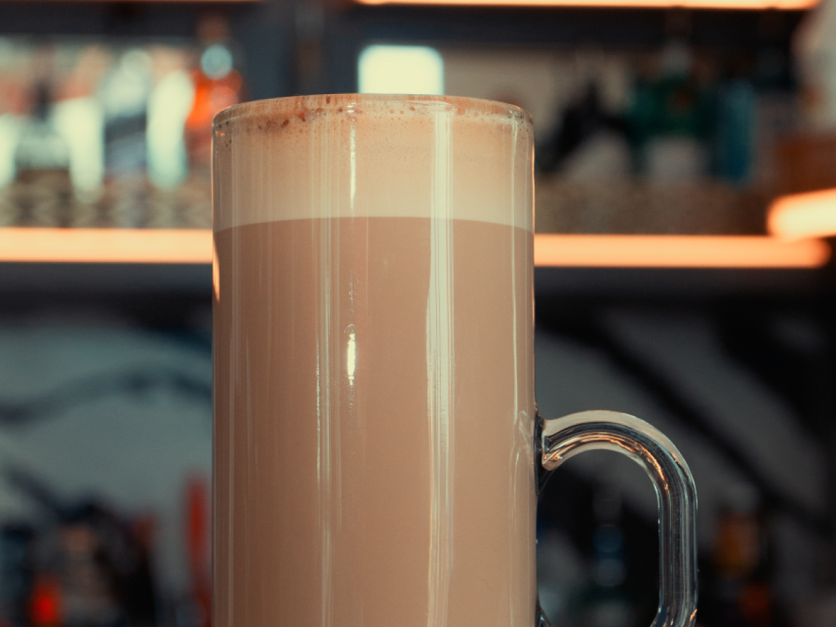 A tall glass mug filled with a layered brown drink, likely hot chocolate or coffee, topped with froth, sits on a counter with blurred background.