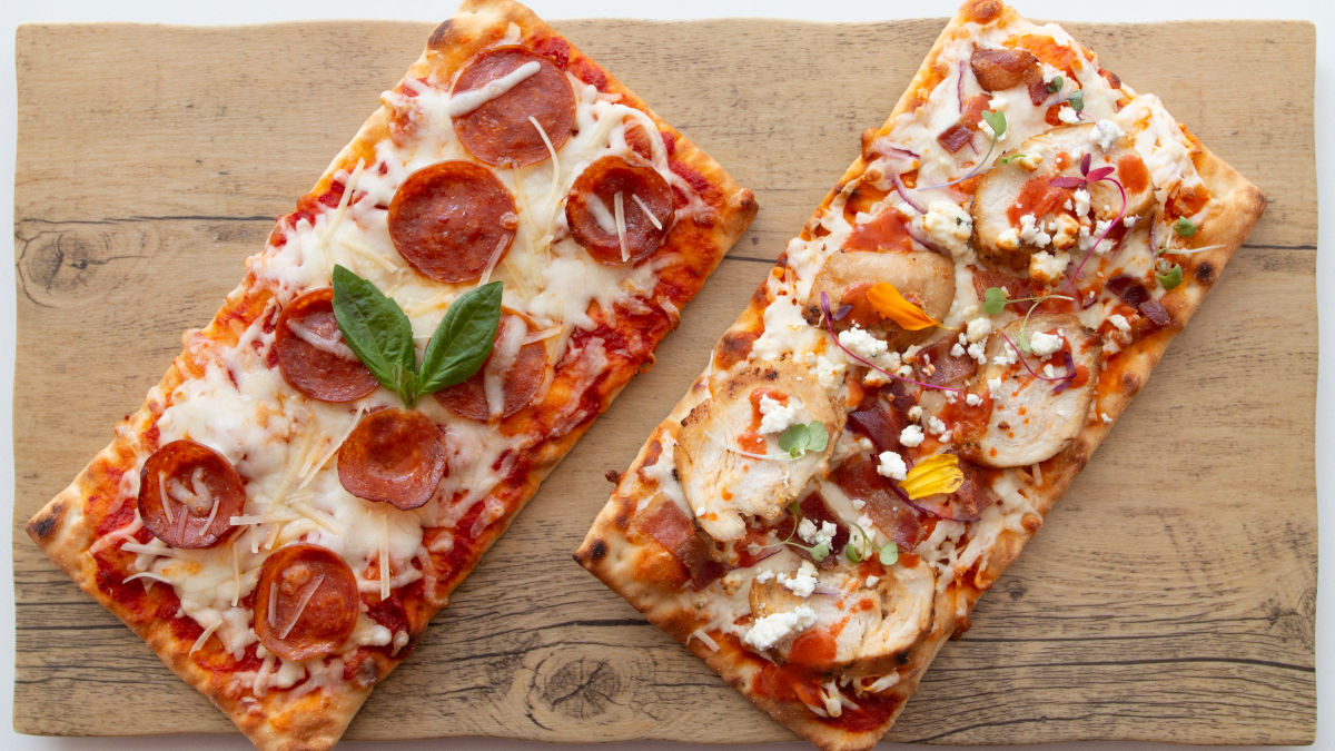 Two rectangular pizzas on a wooden board: one with pepperoni, basil, and cheese; the other with mixed toppings including chicken and vegetables.